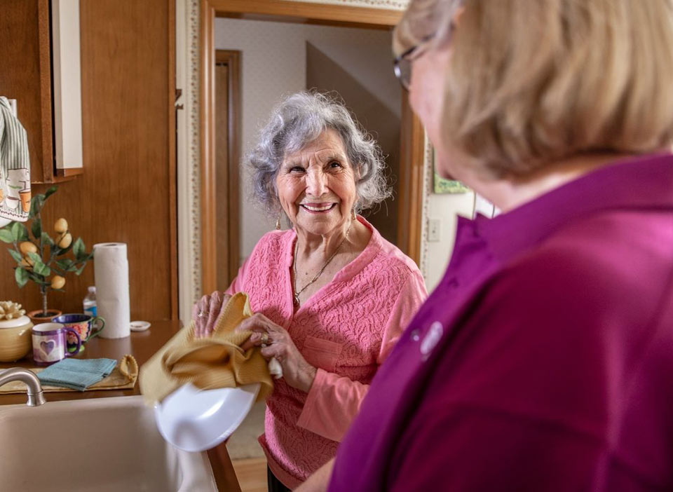 Senior woman and Home Instead Caregiver smiling and washing dishes at home